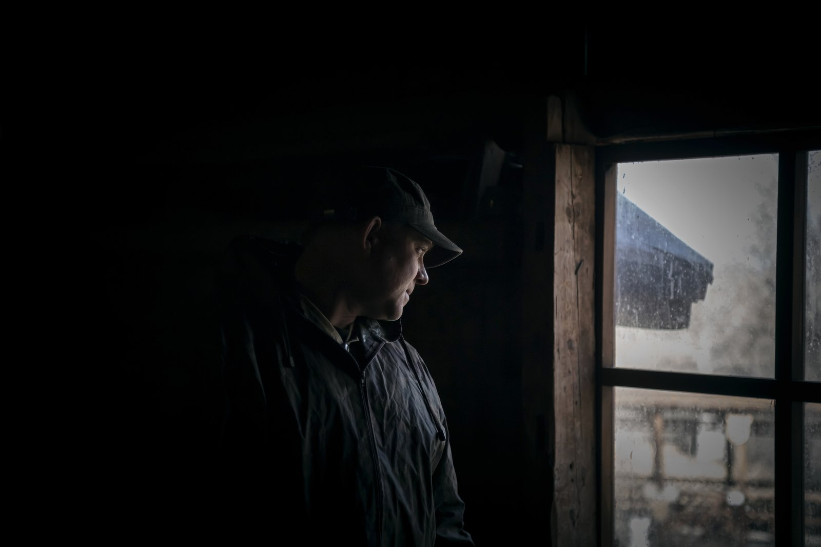 A man stands alone in a dark room looking out a window toward daylight