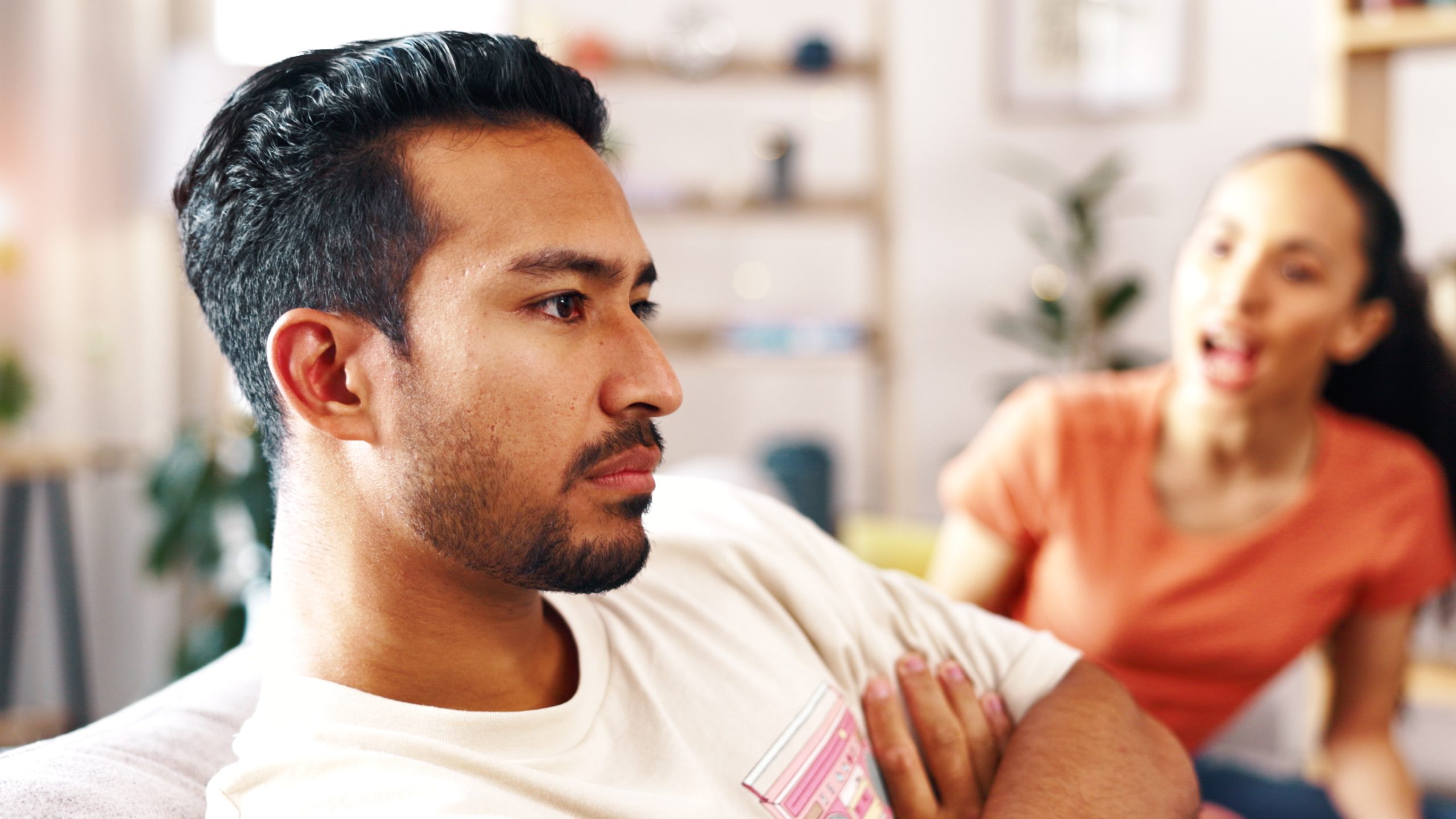 A man sits with his arms crossed, looking away, while his wife speaks emotionally behind him.
