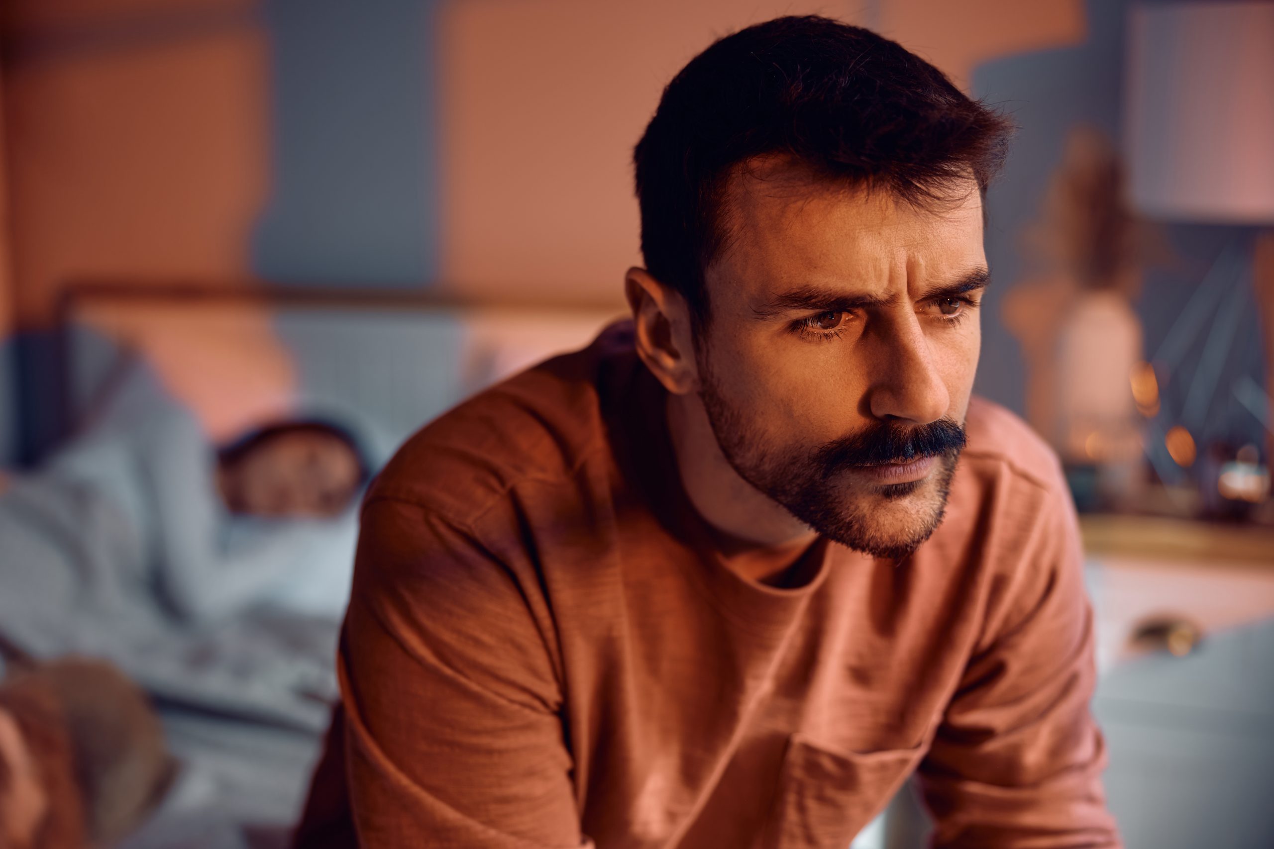 Man with a mustache sitting on the edge of the bed at night, jaw set and staring off, while his partner sleeps behind him